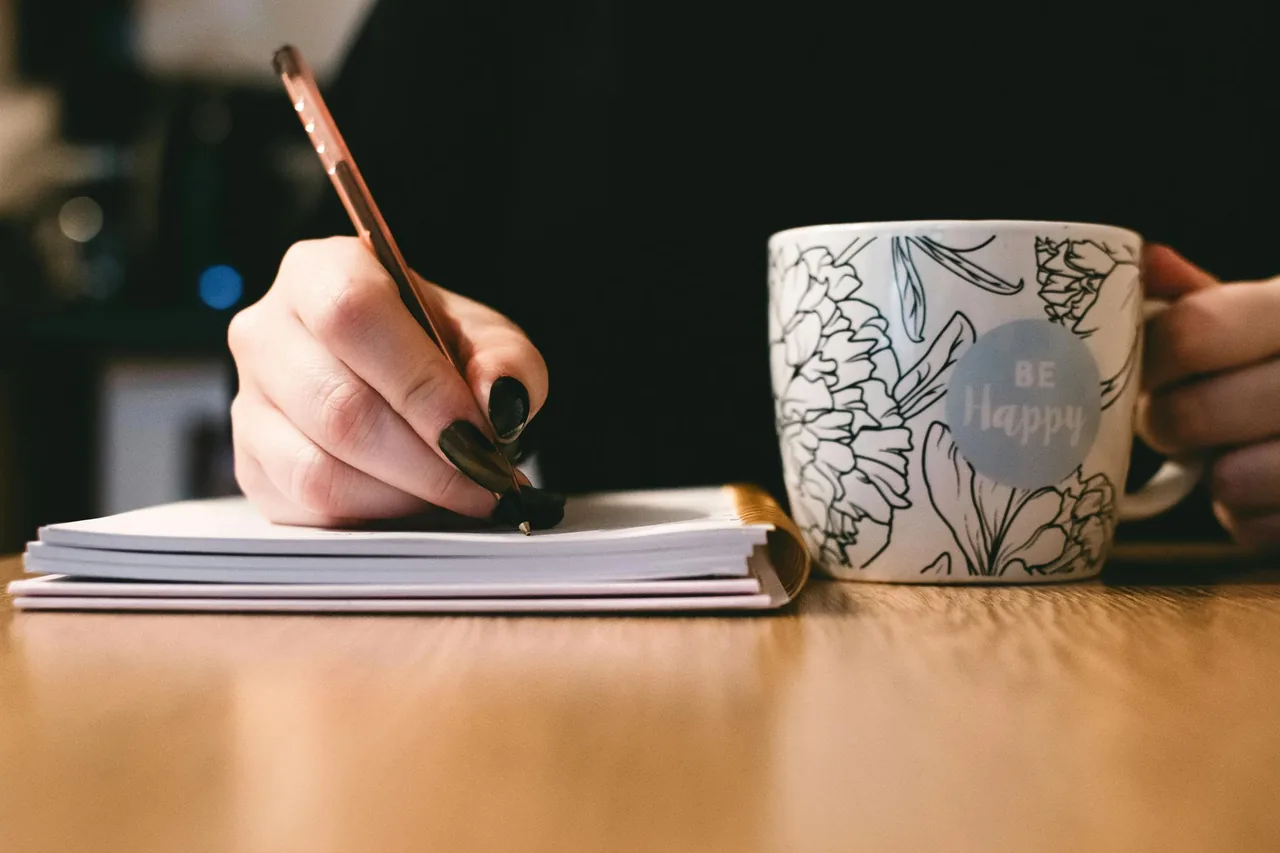Person writing in a notebook beside a mug.
